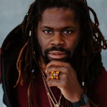 Man with long dreadlocks and a beard poses with his hand on his chin, wearing a maroon shirt, jewelry, a ring, and a black watch against a gray background.