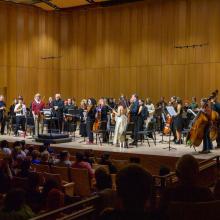 A symphony orchestra stands on stage with instruments, facing an applauding audience in a concert hall with wooden walls.