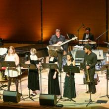 A jazz ensemble performs on stage with vocalists and instrumentalists, including a pianist, drummer, keyboardist, trombonist, and saxophonist, under warm stage lighting.