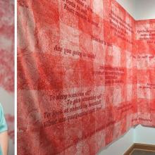A person in a teal top stands beside a red and white textile artwork with text displayed on a wall in a gallery setting.