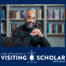 A person with a beard and black turtleneck sits at a table, smiling, in front of a bookshelf. Text reads: “Phi Beta Kappa Society Visiting Scholar Program.”.