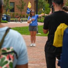 A woman in a blue shirt and shorts leads a group tour outdoors, gesturing with her right hand while people listen and watch.