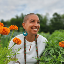A Leah Penniman  wearing a white shirt and beaded necklace, smiles among orange marigold flowers in a green garden.