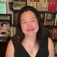 Dr. Chow with long dark hair smiles at the camera, seated in front of bookshelves filled with books and framed photos.