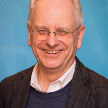 Headshot of a smiling man with short grey hair and glasses wearing a dark blazer over a blue sweater.