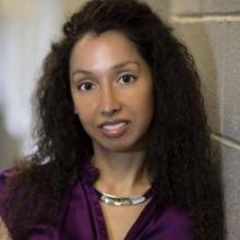 A woman with long, dark, curly hair wearing a purple top and a silver collar necklace.