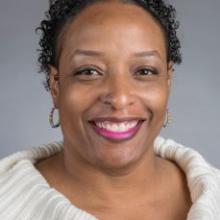 A woman with short curly hair, hoop earrings, and a nose ring smiles at the camera. She is wearing a white sweater and has a neutral gray background.