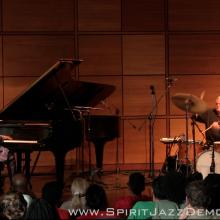 A pianist in a purple jacket and a drummer perform on stage in front of a seated audience; microphones and instruments are visible.