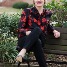 Dr. Neiles sits on a bench wearing a black shirt with red flowers, black pants, and black high heels.