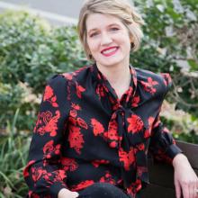 Neiles sits on a bench wearing a black shirt with red flowers, black pants, and black high heels.
