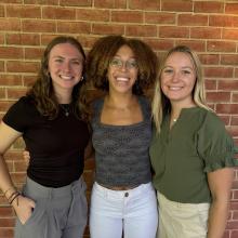 Three women stand outside in front of a brick wall.