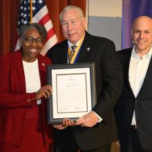 Three people stand together, smiling. The woman on the left is holding a framed document with the man in the center. An American flag and a blue banner are in the background. The man on the right is wearing a suit and has a pin on his lapel.