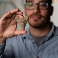 A person wearing glasses and a blue shirt holds up a large fossilized tooth. The tooth is in focus, with the person slightly blurred in the background.