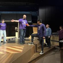 People on stage rehearsing. One person stands on a platform holding a script, while another person reaches out to them. Others watch nearby.