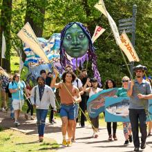 A group of people march outdoors holding banners, flags, a large puppet head, and a cardboard fish, with trees and greenery in the background.
