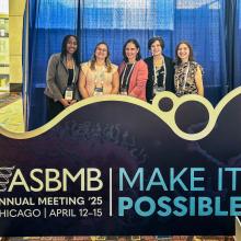 Five people stand behind an ASBMB Annual Meeting 2025 sign in Chicago, featuring the event theme "Make It Possible."