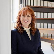 A woman with red hair, wearing a dark blazer and light turtleneck, stands indoors near a window, smiling with hands clasped. A spiral staircase is visible in the background.