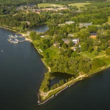 Aerial view of a green, tree-lined waterfront property with several buildings, a marina with boats, and adjacent parking areas along a river or lake.