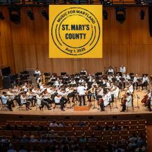 A symphony orchestra performs on stage in a wooden concert hall with a banner overhead reading "Music for Maryland, St. Mary's County, Aug. 1, 2025."