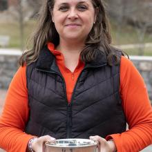 A woman in an orange shirt and black vest stands outdoors, holding a silver bowl with both hands and looking at the camera.