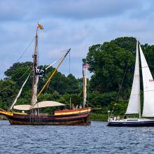 A wooden tall ship with a race committee flag and a modern white sailboat are side by side on a river, with trees and greenery in the background.