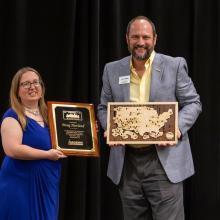 Two people stand in front of a black curtain; one holds a plaque and the other holds a wooden map of the United States, both smiling at the camera.