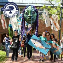 A group of people participate in an outdoor parade carrying banners, a large fish prop, and a green puppet head. A "Best 390 Colleges" Princeton Review badge is overlaid in the top left.