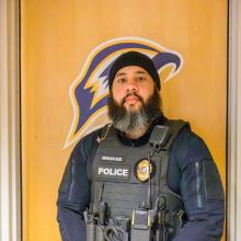 A police officer wearing a tactical vest stands in front of a wooden door with a sports team logo.
