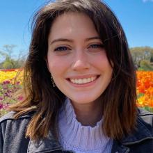 A woman with shoulder-length brown hair smiles outdoors in front of colorful flowers under a blue sky.