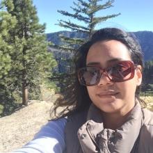 A woman wearing sunglasses and a jacket takes a selfie outdoors with pine trees and mountains in the background.