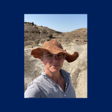 A person wearing a wide-brimmed hat, glasses, and a light shirt stands outdoors on a dusty path with rocky terrain in the background under a clear sky.