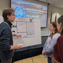 A student presents a research poster to two women with a display screen and American flags visible in the background.