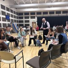 A group of students sits in a circle in a classroom while the instructor (Villada) stands and speaks; another person stands nearby and takes a photo with their phone.