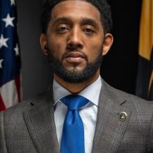 A man in a plaid suit jacket and blue tie stands in front of American and state flags, facing the camera with a neutral expression.