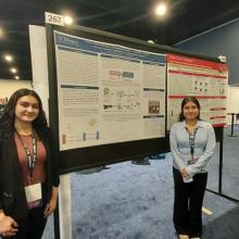 Two women stand beside a large scientific poster display at a conference with other posters and attendees visible in the background.