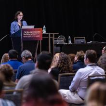 A woman speaks at a podium during a conference, addressing an audience seated in rows. A sign on the podium reads "ASBMB Make It Possible.