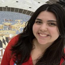 A young woman with long dark hair smiles at the camera, standing indoors on a marble floor featuring a decorative crest design.