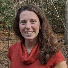 A woman with long brown hair, wearing a red cowl-neck top, stands outdoors in a wooded area with fallen leaves on the ground.