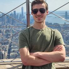 A man in sunglasses and a green shirt stands with arms crossed on an observation deck, with a city skyline and skyscrapers in the background.