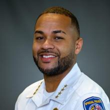A man in a white police uniform with gold stars on his collar and a "Special Police" patch on his shoulder, smiling in front of a gray background.