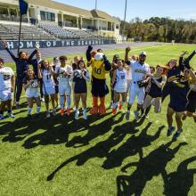 A group of athletes in sports uniforms and a mascot jump and pose for a group photo on a turf field in front of a stadium building.