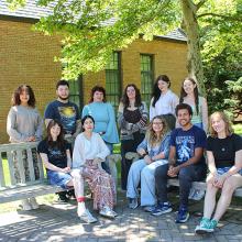 A group of eleven people pose for a photo outside on benches in front of a brick building and trees on a sunny day.