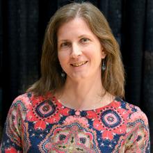 A woman with long brown hair wearing a red and blue patterned top stands in front of a black curtain, smiling at the camera.