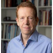 A man with short red hair wearing a blue shirt is standing in front of bookshelves filled with books. He is looking directly at the camera with a neutral expression.