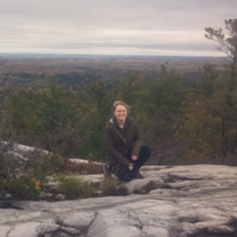 Morgan Siewert kneeling on a stone cliff overlooking an evergreen forest