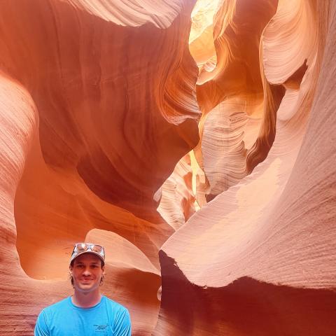 Matthew, wearing blue long sleeve shirt and hat, smiling and standing inside a slot canyon in Northern Arizona.