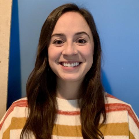 Rachel Heiss is smiling at the camera, wearing an orange, yellow and white striped sweater and standing in front of a blue wall.