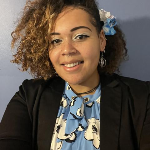 Headshot photo of Aris Nazarova wearing a blue shirt with flowers, a black blazer and has a blue flower in her hair.