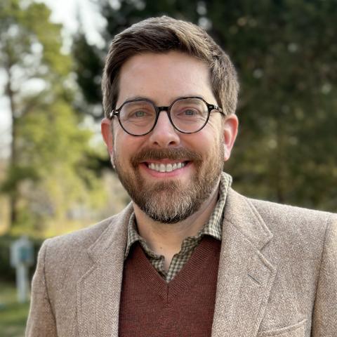 Head-and-shoulders image of Brian C. Smithson wearing a tan blazer over an orange sweater and grey, button-down shirt. He is a white man with short brown hair, brown glasses, and a brown beard.