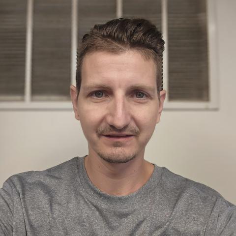 Darin, a white man with dirty blonde short hair, is inside a lit room, wearing a grey smcm physical plant staff shirt.